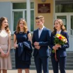 Skupina tínedžerov v elegantnom oblečení pred školou na slnečný deň s kvetmi a úsmevom.